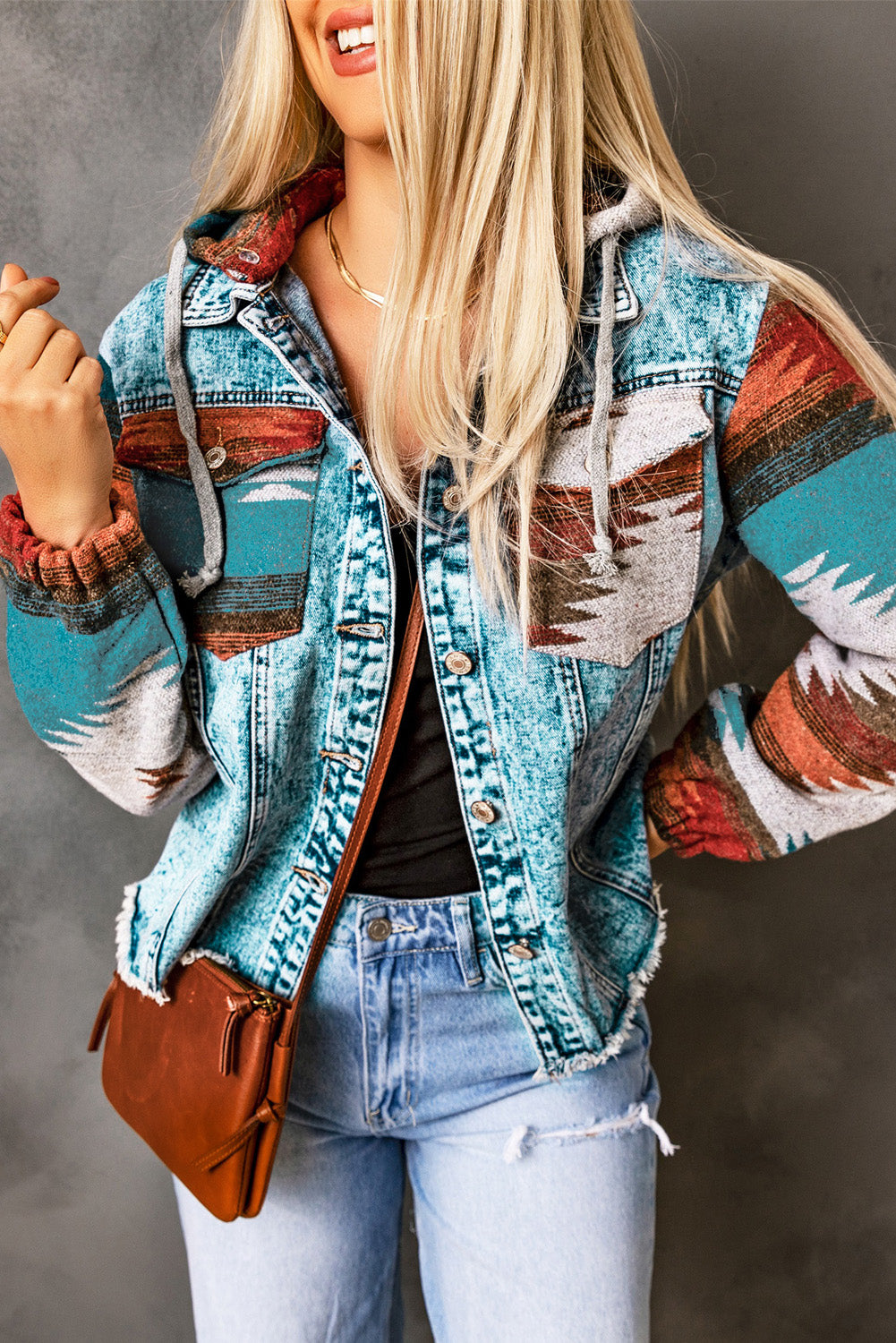 Woman wearing an Aztec print denim jacket with frayed hem and colorful sleeves, styled with jeans and a brown crossbody bag”